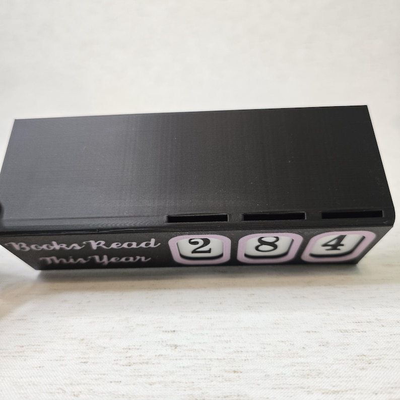 May include: A black rectangular book counter with the text "Books Read This Year". The counter displays the number "284" in white digits on a light purple background. The item is designed to track the number of books read.