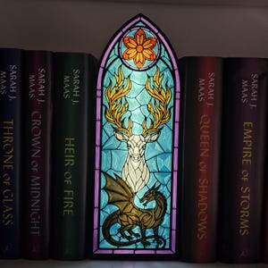 May include: A rectangular stained glass-style lamp with an arched top, featuring a white stag with golden antlers and a golden dragon below it. The background is light blue with a geometric pattern and an orange flower at the top, framed in purple.