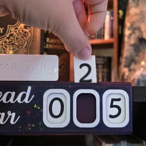 May include: A black and white Tome Tally reading tracker with the words "Read Year" in white script. A hand is placing the number "2" into the tracker, which currently reads "005". A white rectangular block with the words "Tome Tally" is also visible.