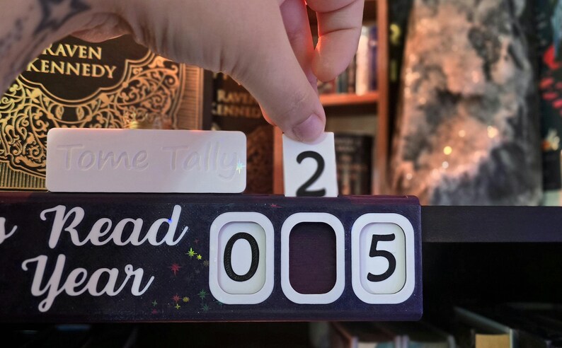 May include: A black and white Tome Tally reading tracker with the words "Read Year" in white script. A hand is placing the number "2" into the tracker, which currently reads "005". A white rectangular block with the words "Tome Tally" is also visible.