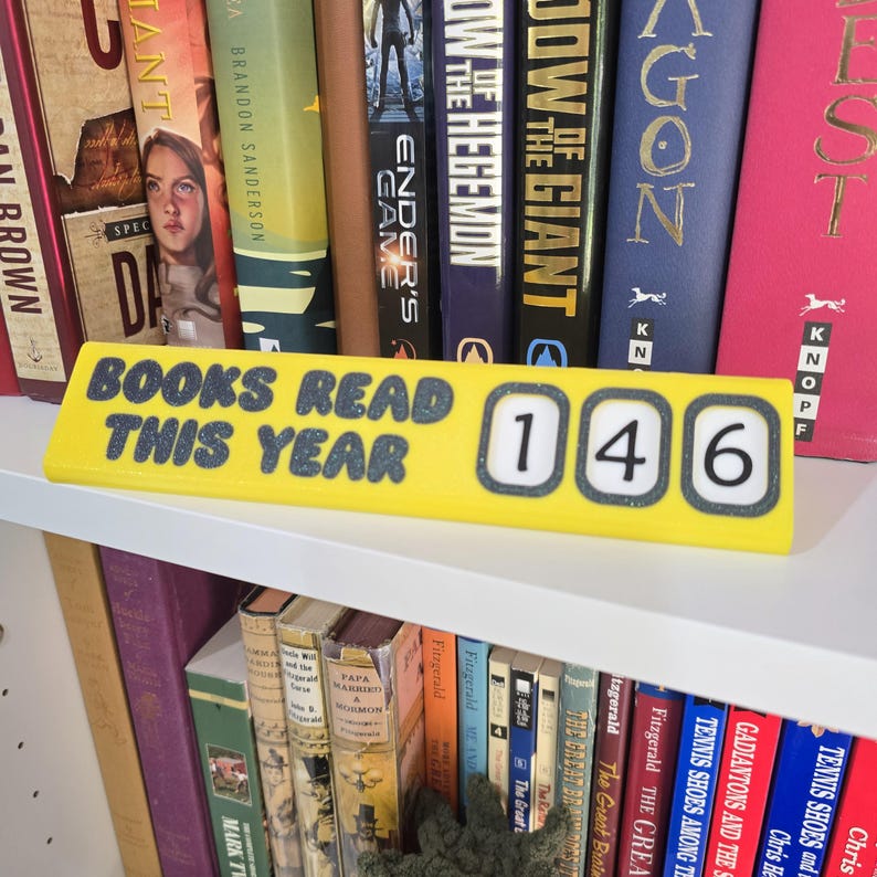 May include: A yellow rectangular book tracker with the text "BOOKS READ THIS YEAR" and the number "146". The tracker is placed on a white shelf in front of a row of books.
