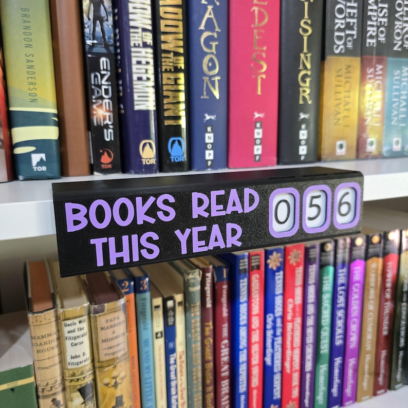 May include: A black counter displays "BOOKS READ THIS YEAR" in purple lettering, with a reading count of 056. The counter sits in front of a shelf filled with books of various colors and sizes, creating a library theme.