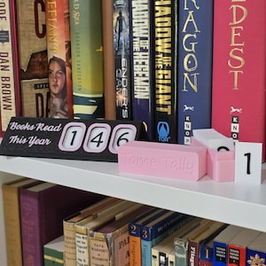 May include: A black and pink book counter with the text "Books Read This Year" and the number "146". A pink "Tome Tally" block and white number tiles are also present. The items are on a white shelf with books in the background.