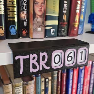 Può includere: Un organizer per libri nero montato su mensola con la scritta "TBR 061" in rosa. L'organizer è progettato per contenere libri ed è posizionato su una mensola bianca, con una collezione di libri visibile sullo sfondo.