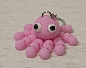 Könnte beinhalten: Ein rosa Tintenfisch-Schlüsselanhänger mit schwarzen Augen. Der Tintenfisch hat acht Tentakel und ist aus einem weichen, biegsamen Material gefertigt.