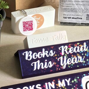 May include: A collection of book-themed decor items. Includes a white rectangular block with "ary Tally" and a QR code tag, a "Tome Tally" block, and two dark blue bookends with the text "Books Read This Year" and "Books In My Library".