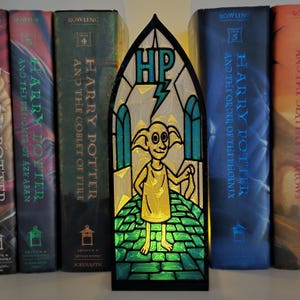 May include: A stained glass-style night light featuring the character Dobby from the Harry Potter series. The light has a black frame and a green, yellow, and blue color scheme. The letters "HP" and a lightning bolt are at the top.