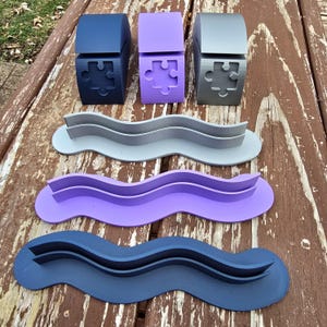 May include: Three puzzle-shaped boxes in navy, lavender, and gray, each with a matching wavy tray. The boxes have a puzzle piece cutout. The trays are designed with a wavy shape to hold items.