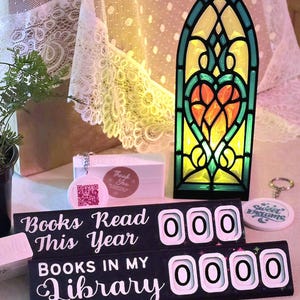 May include: A decorative stained glass-style light with a heart design, a book counter, and a keychain. The book counter reads "Books Read This Year" and "Books in My Library". The color scheme includes black, green, yellow, and orange.