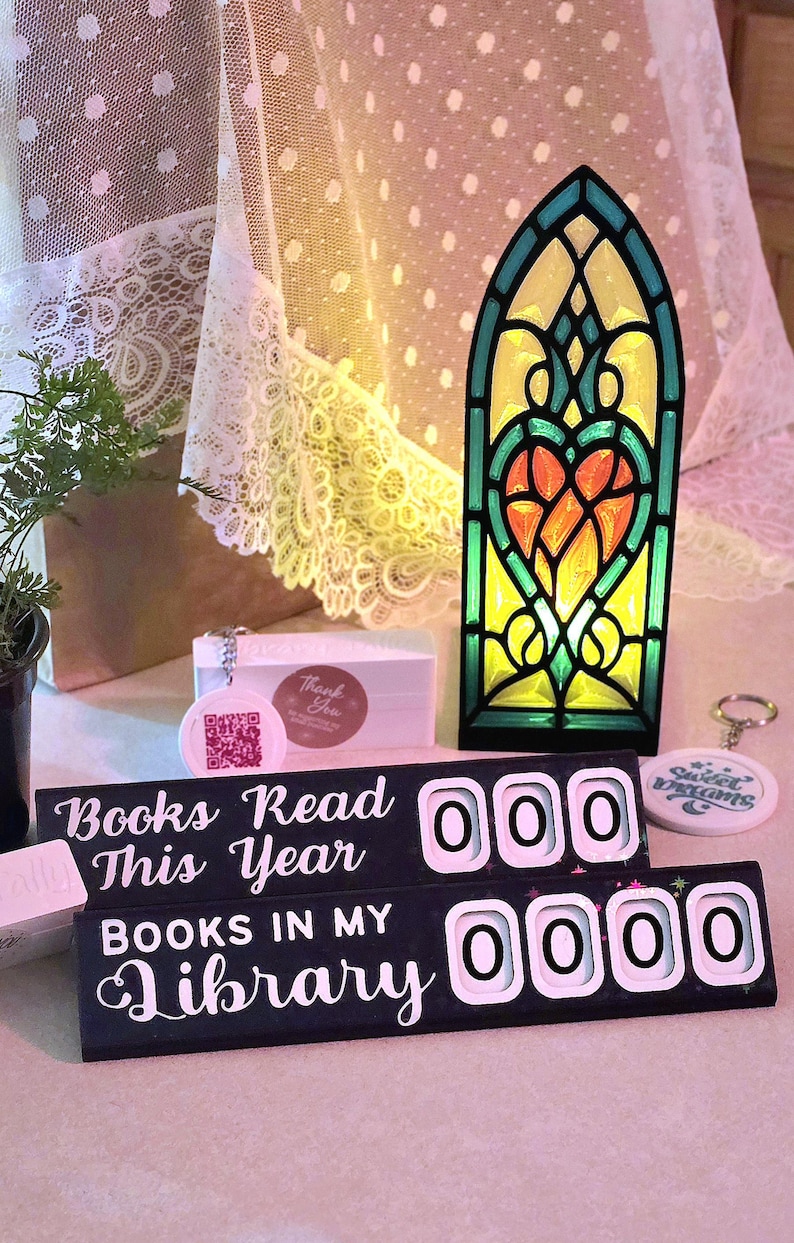 May include: A decorative stained glass-style light with a heart design, a book counter, and a keychain. The book counter reads "Books Read This Year" and "Books in My Library". The color scheme includes black, green, yellow, and orange.