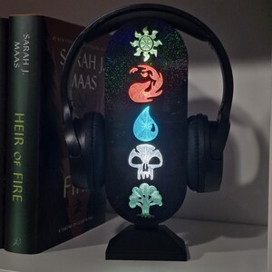 May include: A black headphone stand with a vertical oval display featuring five glowing symbols in a column: a sun, a flame, a water drop, a skull, and a tree. The stand has a glittery finish and supports a pair of black over-ear headphones.