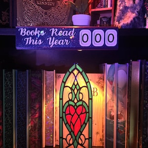 May include: A decorative shelf with books, a stained glass heart-shaped lamp, and a sign that says "Books Read This Year 000." The books have gold lettering and are in a variety of colors. A red flower sits on the shelf.