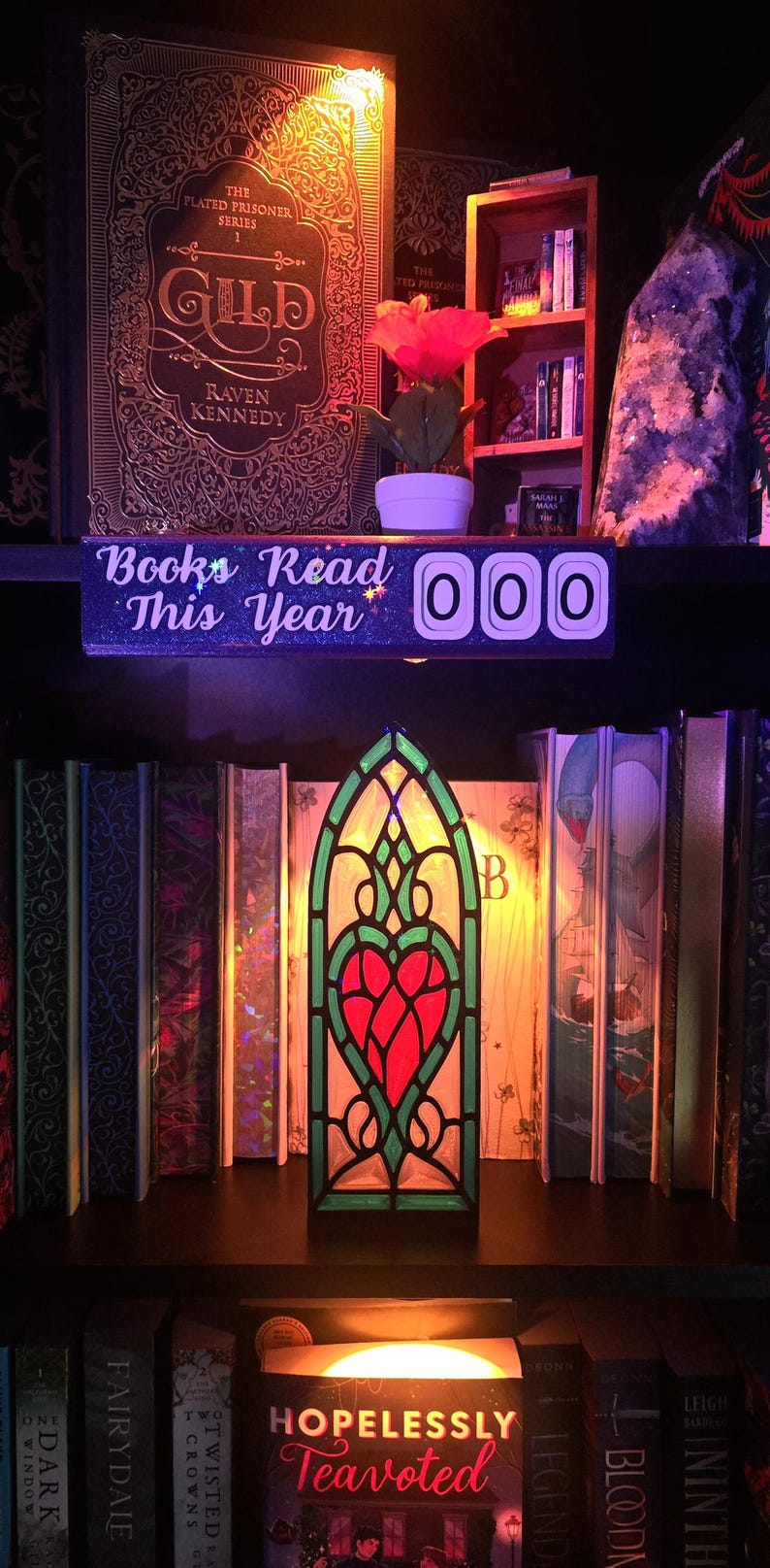 May include: A decorative shelf with books, a stained glass heart-shaped lamp, and a sign that says "Books Read This Year 000." The books have gold lettering and are in a variety of colors. A red flower sits on the shelf.