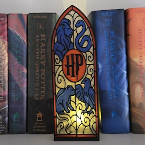 Könnte beinhalten: Ein Nachtlicht im Buntglasstil mit dem Harry Potter-Logo und den Hausmaskottchen. Das Design zeigt einen Löwen, eine Schlange, einen Bären und einen Adler in Blau, mit einem orangefarbenen und schwarzen HP-Emblem. Das Licht steht vor Harry Potter-Büchern.