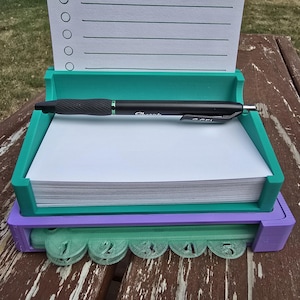 Könnte beinhalten: Ein mehrfarbiger Schreibtisch-Organizer mit Notizblock, Stift und nummerierten Registerkarten. Der Organizer besteht aus türkis, lila und grünem Kunststoff. Ein schwarzer Stift liegt auf dem Notizblock. Der Notizblock hat eine Liste von Kreisen und Linien.