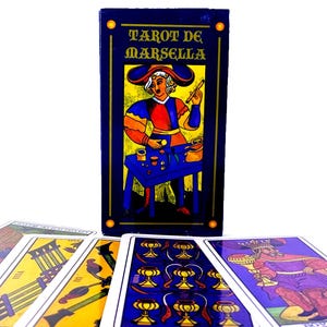 May include: A box of Tarot de Marseille playing cards with a blue background and gold trim. The box features a colorful illustration of a man in a blue and red costume holding a wand. The cards are spread out on a white surface.