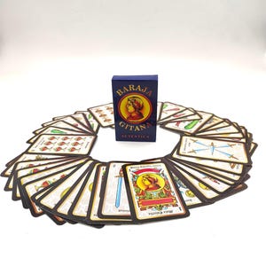 May include: A deck of Baraja Gitana Autentica playing cards arranged in a circular fan pattern around a central blue box. The cards feature colorful illustrations of swords, cups, and figures with text including Mala Estrella.