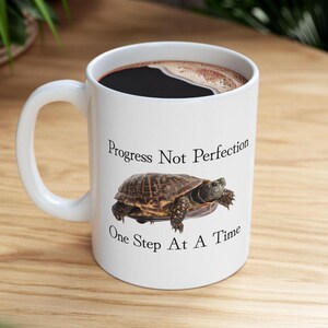 Può includere: Una tazza da caffè in ceramica bianca con un manico nero. La tazza presenta una grafica di tartaruga marrone e nera e il testo "Progress Not Perfection One Step At A Time".