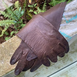 May include: A pair of dark brown leather gloves with a smooth, slightly textured finish and a classic cut. The gloves feature stitched detailing along the fingers and a wide cuff that extends past the wrist, showcasing a natural grain pattern.