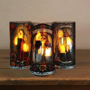 May include: Three decorative glass candle holders with colorful religious and Day of the Dead themed designs. Each cylindrical holder features a different image, including a saint, a skull, and an angel. The candle holders are illuminated from within, creating a warm glow.