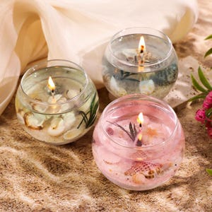 Peut inclure: Trois bougies rondes en verre allumées sur le thème de la plage. Chaque bougie contient du sable, des coquillages et de la verdure. Une bougie a de la cire rose, tandis que les autres ont de la cire transparente. Les bougies sont posées sur une surface sablonneuse.