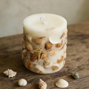 May include: A cylindrical candle decorated with various seashells and a starfish. The candle is off-white, with the seashells in shades of brown and tan. Several seashells are scattered around the candle on a wooden surface.