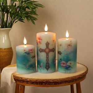 May include: Three pillar candles with a floral and butterfly design. The center candle features a cross. The candles have a blue and white ombre effect with a flickering flame. The candles are on a small, round wooden table.
