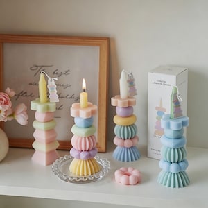 May include: A collection of pastel-colored geometric candles. One candle is lit, and the others are unlit. The candles are stacked with various shapes, including flowers, spheres, and cylinders. A small, pink, flower-shaped candle sits in front.