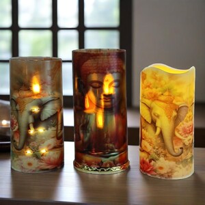 May include: Three decorative LED candles with Buddha and elephant designs. The candles are cylindrical, with warm yellow and orange tones. The designs include floral and lotus patterns, with a flickering light effect.