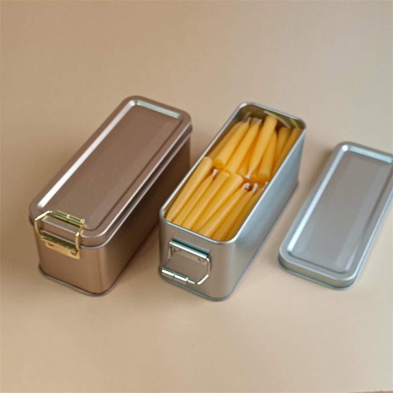 May include: Two rectangular metal tins with lids. One tin is silver and contains a stack of yellow beeswax candles. The other tin is gold and has a gold latch.