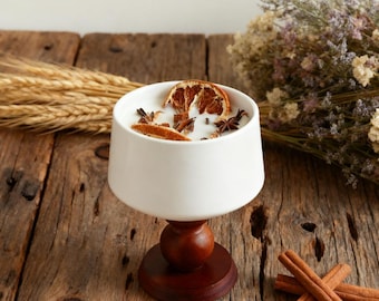 May include: A white candle in a pedestal-style holder, decorated with dried orange slices and star anise. The candle sits on a dark wooden base, with cinnamon sticks nearby. The overall theme is a cozy, aromatic home decor item.