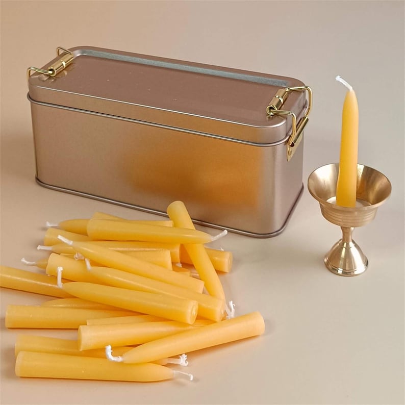 May include: A set of 20 small, yellow beeswax candles in a gold metal tin with a hinged lid. The candles are arranged in a pile and there is a small brass candle holder with a single yellow beeswax candle.