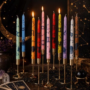 May include: A collection of ten tall, slender taper candles in various colours, each featuring dark blue zodiac signs and constellation patterns. The candles are lit and held upright in individual gold-toned metal stands on a dark surface.