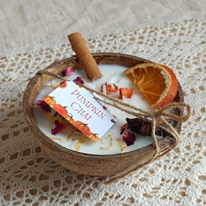 Może przedstawiać: Świeca o zapachu pumpkin chai w skorupie kokosa. Świeca jest ozdobiona laską cynamonu, suszonym plasterkiem pomarańczy i suszonymi kwiatami. Mała etykieta głosi "Pumpkin Chai". Świeca jest związana sznurkiem.