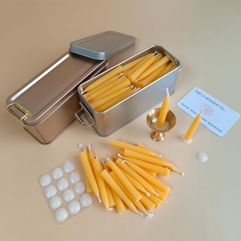 May include: A set of small, yellow beeswax candles in a silver tin with a lid. The candles are arranged in rows and are ready to be used. The tin is sitting on a light brown surface. There are also a few white candle holders and a small, gold candle holder with a lit candle. A card with the text "Take a moment to... Breathe, Relax, Renew" is also visible.