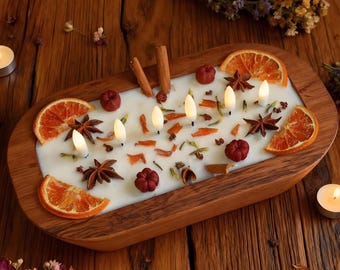 Könnte beinhalten: Ein rechteckiger Kerzenhalter aus Holz mit einer weißen Kerzenbasis, verziert mit Orangenscheiben, Sternanis und kleinen roten Kürbissen. Die Kerze hat fünf kleine, leuchtende, weiße LED-Lichter. Zimtstangen und getrocknete Kräuter sind ebenfalls enthalten.