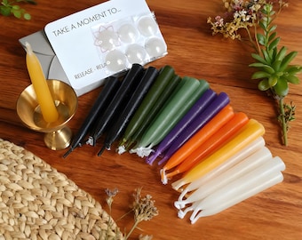 May include: A collection of colorful taper candles in various shades, including black, green, purple, orange, yellow, and white. A small brass candle holder with a yellow candle is on the left. A card with the text "TAKE A MOMENT TO... RELEASE - RELAX" is also present.