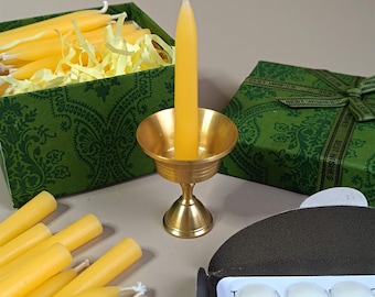 Può includere: Un set di candele di cera d'api gialle in una scatola verde con un portacandele in metallo dorato. La scatola ha un nastro verde con le parole "Auguri" stampate su di esso. Le candele sono disposte in una scatola con carta triturata gialla.