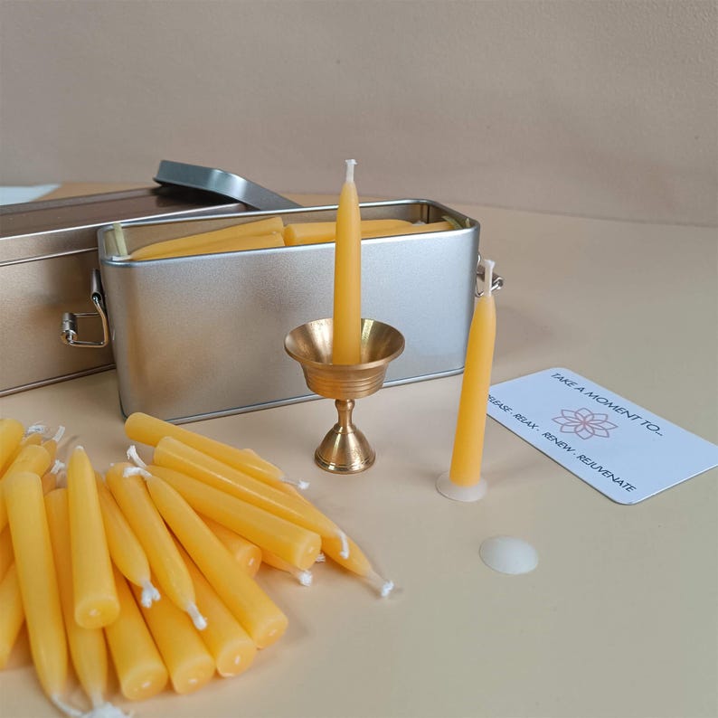 May include: A set of small yellow beeswax candles in a silver tin with a brass candle holder. The candles are arranged in a pile and there is a card that says "Take a moment to..." with a flower design.