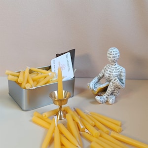May include: A tin box filled with small yellow beeswax candles, a single candle in a brass candlestick, and a small card that reads "Take a moment to..." with a floral design.  There is also a small figurine of a person reading a book made of paper with the word "books" printed all over it.