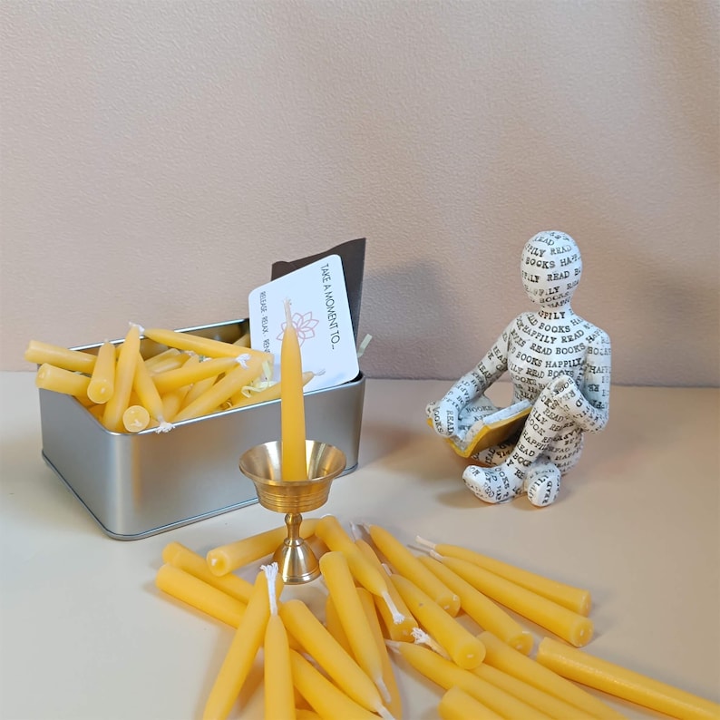 May include: A tin box filled with small yellow beeswax candles, a single candle in a brass candlestick, and a small card that reads "Take a moment to..." with a floral design.  There is also a small figurine of a person reading a book made of paper with the word "books" printed all over it.