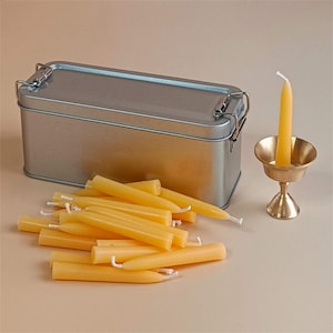 May include: A silver metal tin with a hinged lid containing a set of small, yellow beeswax candles. A single candle is displayed in a small, gold metal candle holder.