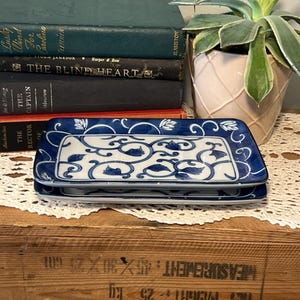 May include: Two rectangular ceramic dishes with a blue and white floral design. The dishes have a white centre with blue floral patterns and a blue border. They are stacked on a wooden surface, with books and a plant in the background.