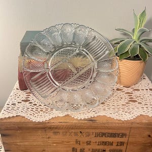 May include: Clear glass egg serving platter with scalloped edges and multiple compartments. The center features a diamond pattern, and the outer edge has egg-shaped indentations. A small potted plant sits to the side.