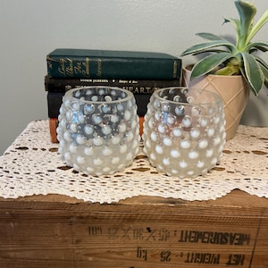 May include: Two clear glass votive candle holders with white, raised, bubble-like designs. The candle holders sit on a white lace doily, with books and a potted plant in the background. The wooden crate below has text.