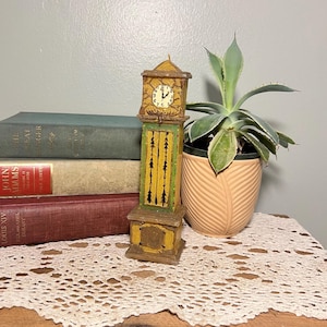 Puede incluir: Una figura en miniatura de reloj de abuelo pintada a mano. El reloj presenta una combinación de colores amarillo y verde con detalles en negro. La esfera del reloj muestra la hora. La figura está colocada sobre una superficie de madera con libros y una planta en maceta en el fondo.
