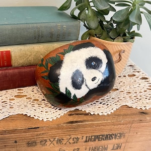 May include: Hand-painted panda bear rock with a white face, black eyes, and black ears. The panda is surrounded by green leaves and rests on a brown rock. The rock is on a white lace doily.