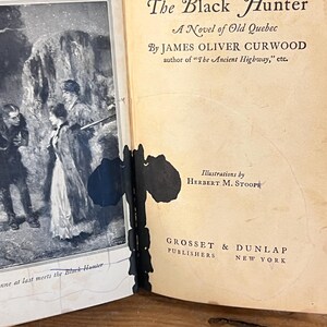 K&ouml;nnte beinhalten: Aufgeschlagenes Buch mit dem Titel "The Black Hunter" von James Oliver Curwood. Das Cover zeigt eine Schwarz-Wei&szlig;-Illustration von Personen in einer verschneiten Landschaft. Herausgegeben von Grosset & Dunlap, New York.