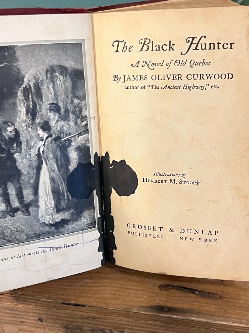 K&ouml;nnte beinhalten: Aufgeschlagenes Buch mit dem Titel "The Black Hunter" von James Oliver Curwood. Das Cover zeigt eine Schwarz-Wei&szlig;-Illustration von Personen in einer verschneiten Landschaft. Herausgegeben von Grosset & Dunlap, New York.
