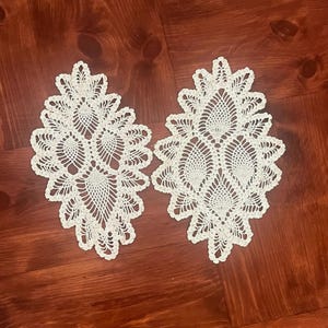 May include: Two white, oval-shaped, crocheted doilies. Each doily features a detailed, openwork design with a central motif and a scalloped edge. The doilies are displayed on a dark wood surface.